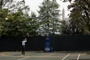 President Obama shoots a basketball into a hoop. He is dressed professionally and is on the White House Basketball court. A White House logo is on the base of the basketball hoop.