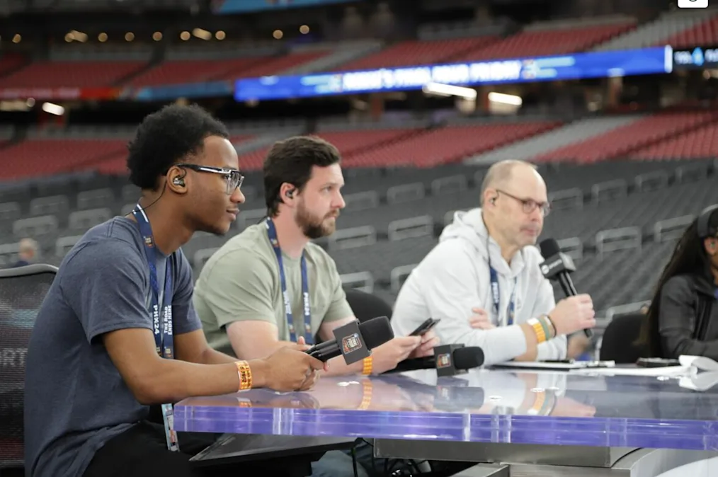 A young Black man with a dark skin tone sits behind an anchor desk with TNT Sports at the NCAA Division I Men’s Basketball Final Four in Phoenix
