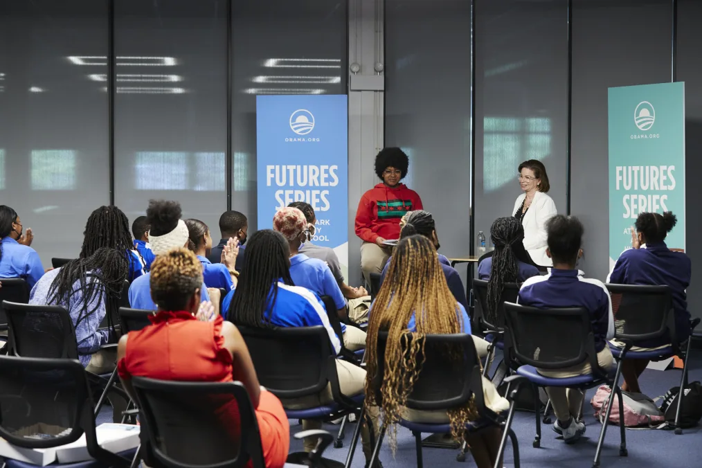 A group of students in matching blue jackest partake in a furture series meeting hosted by the Obama Foundation. The leaders of the talk are a medium-deep toned woman with an afro and glasses wearing casual attire seated next to a older light skin toned woman with medium length brown hair, glasses, and a white blazer.