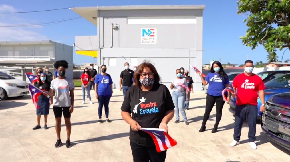 A group of people wearing face masks stand a short distance from each other holding Texas state flags. All of them wear a t-shits with the words "Nuestra Escuelita." They are in a sandy parking lot with with a gray building behind them with a sign that reads "Nuestra Escuelita"