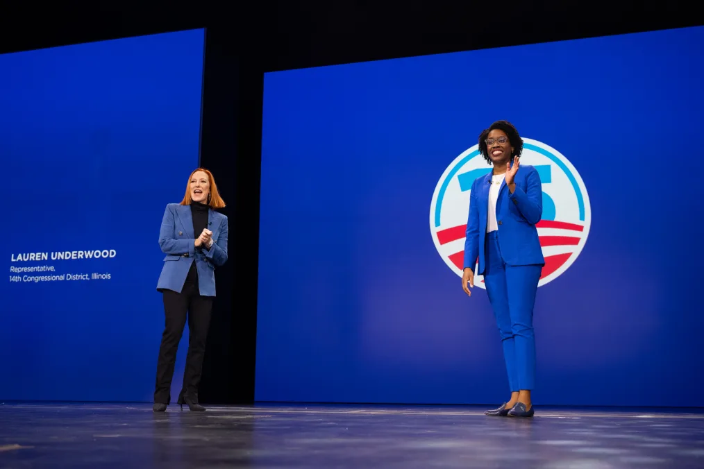 Two women are standing on a stage in an indoor auditorium. Behind the two women is a blue wall, split in two: on one side of the wall is the Obama 15th anniversary logo. The logo has a big number 15 in the center in the color light blue. In the logo are red stripes. On the other side of the wall is the bio of Representative Lauren Underwood that reads, “Lauren Underwood, Representative, .” The first woman in the picture is Jen Psaki: she has a pale complexion and bright red hair cut into a bob. She is smiling and clapping her hands together. She is wearing a blue blazer with gold buttons on top of a black turtleneck, black pants, and black shoes with a thin heel. The second woman in the photo is Representative Lauren Underwood: she has brown skin, dark curly hair that lands at the nape of her neck, and lucite/clear glasses. She is smiling and waving to the crowd. She is wearing a royal blue blazer and pants with a white blouse underneath, and black leather loafers on her feet. 
