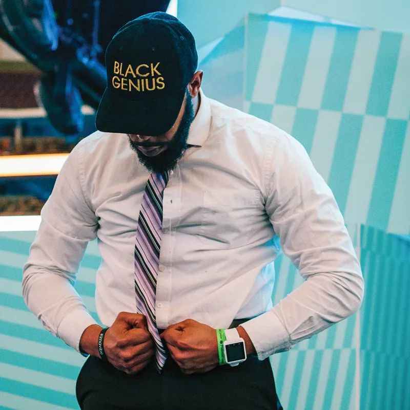 A young man wearing a collared shirt and tie, and a hat that reads "Black Genius" looks down with his fists clenched during the My Brother's Keeper Alliance Youth Opportunity Summit in Newark, New Jersey.