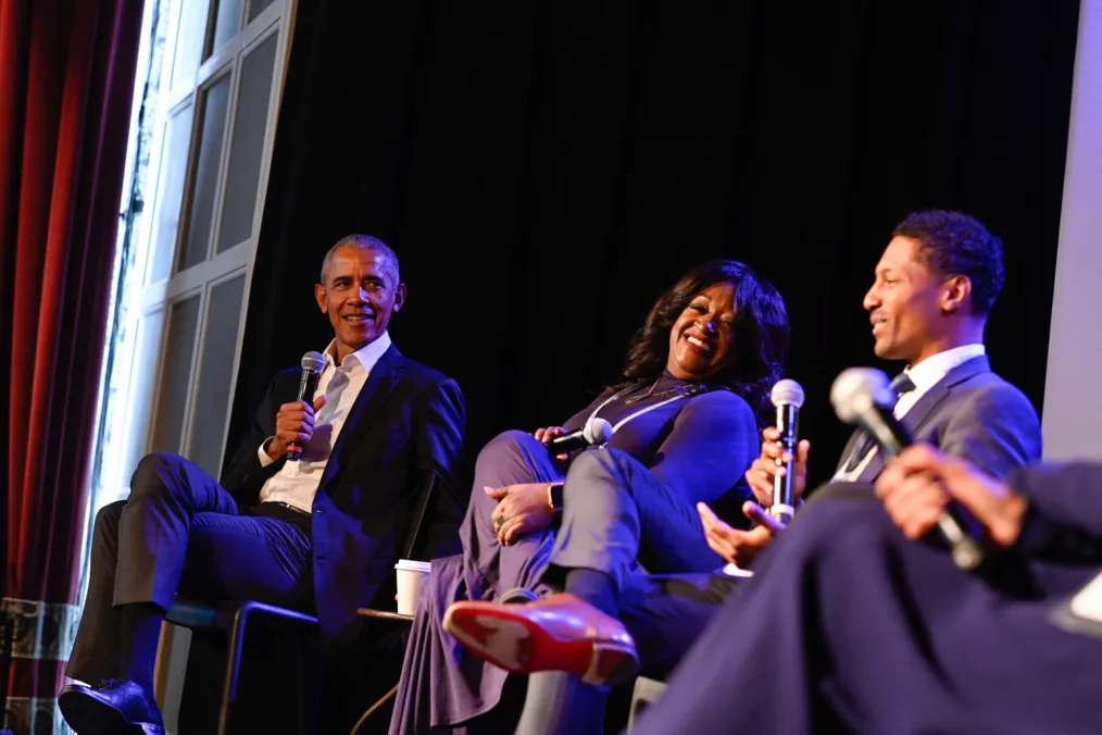 President Obama sits on stage holding a microphone and looking right at two people of varying skin tones and clothing, also seated.