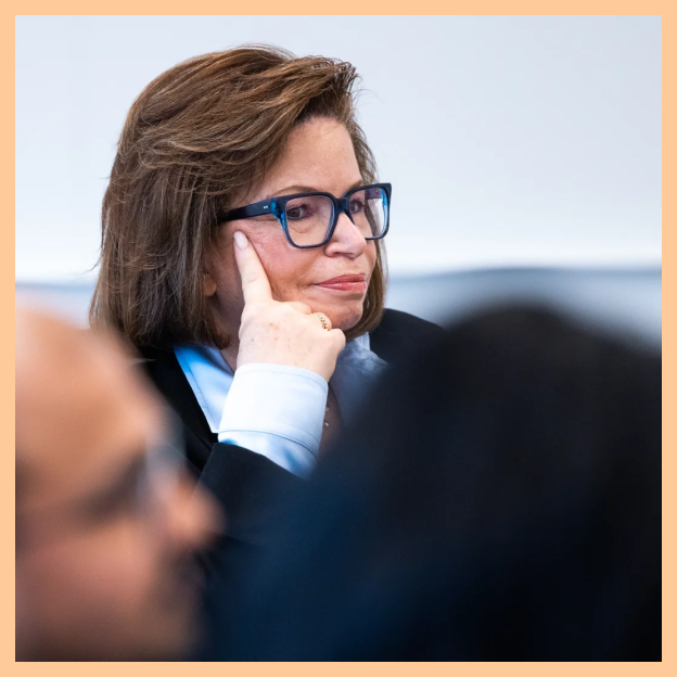 Valerie Jarrett sits listening with her hand up to her face. She wears blue and black glasses, a light blue blouse and a black jacket.