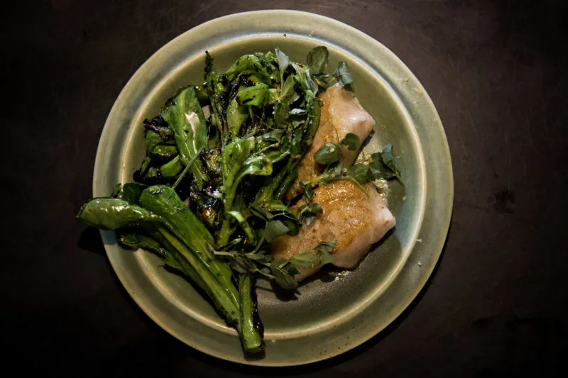 A light green ceramic plate with cooked greens to the left and two pieces of lightly browned meat to the right. The plate is atop a dark wood surface. 