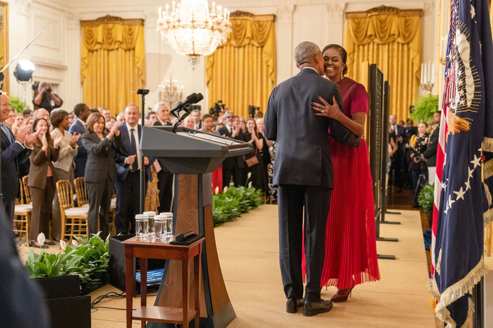President Barack Obama and First Lady Michelle Obama hug on stage in front of a crowd of people with a variety of skin tones. 