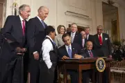 Barack Obama a man with golden brown skin sitting at a table wearing a formal suit while writing with a diverse group of people behind him with mainly neutral light skin and a little boy with brown skin wearing a formal suit looking at the paper Barack is writing on 