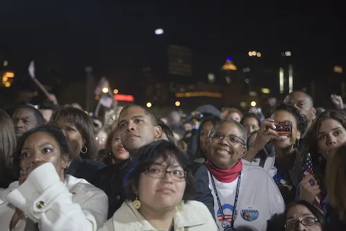 A crowd of people look at something not shown in the photo. Some seem happy, some surprised and others unmoved. A city skyline is lit up behind them in the dark. 