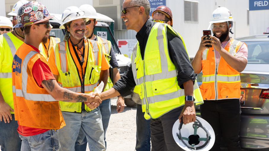 Barack Obama shakes hands with construction workers wearing neon yellow vests and hardhats. One man on the right takes a photo with his cell phone.
