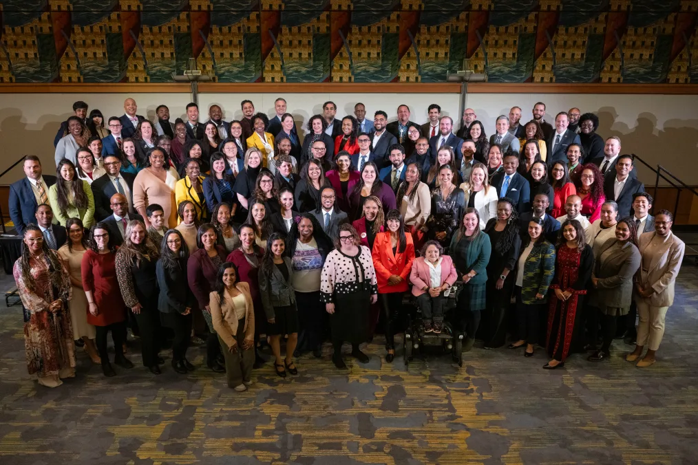 A large group of people with a range of light to dark skin tones pose for a photo. All are dressed professionally. 