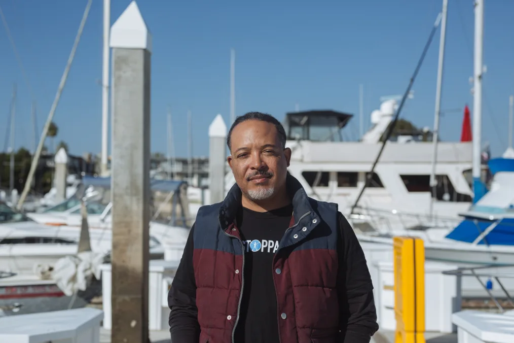 A man with a medium skin tone is shown outside in front of boats looking forward toward the camera.