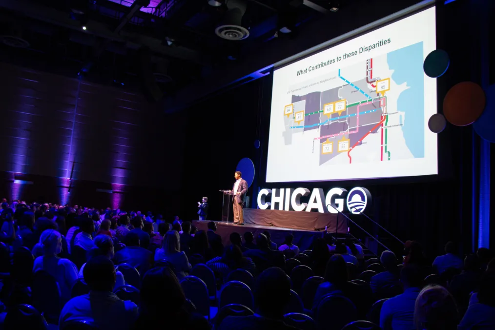 A man with a deep medium skin tone stands on stage in a spotlight in a front of a large sign that reads " Chicago" and a large white screen with an image of a map and text that reads "What Contribute To These Disparities". A group of people sit in chairs in a dark blue lighting 