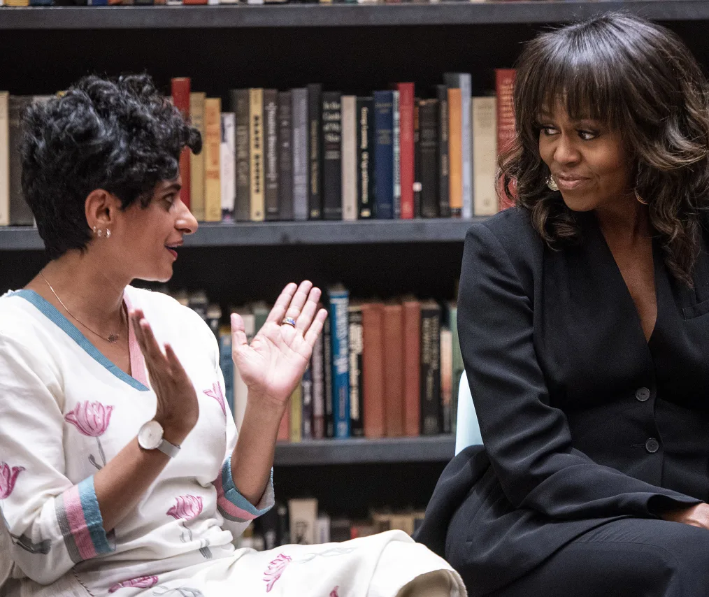 A woman wearing a floral-patterned dress has her hands raised in a "V" shape and looks at Michelle Obama who is partially cut out of the photo. Behind them are shelves of books.