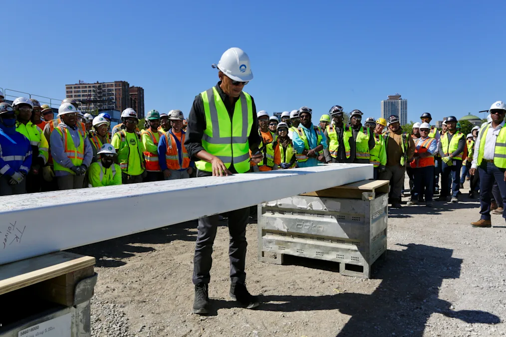 On a clear blue-sky day, President Obama stands in a hard hat and a brightly colored safety vest at the Obama Presidential construction site and signs a beam. A large group of construction workers with various skin tones also wearing safety clothing surround him. 