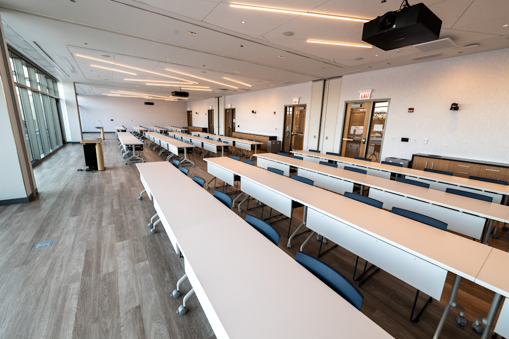 In a large classroom with light wood floors and white walls, long white tables with blue chairs are arranged in neat rows. Two black podiums sit near large floor-to-ceiling windows on the left. Projectors hang from the high white ceiling, and two exit doors are on the right back wall.