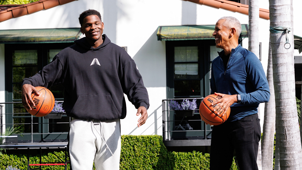 Anthony Edwards and Barack Obama stand next to each other outdoors. Each man is holding a NBA Spalding basketball in their hands. Anthony has a deep skin complexion short coiled hair and a goatee. He is wearing a black sweatshirt. Barack Obama has medium deep skin tone and closely cropped gray hair. He is wearing a light colored button down shirt.