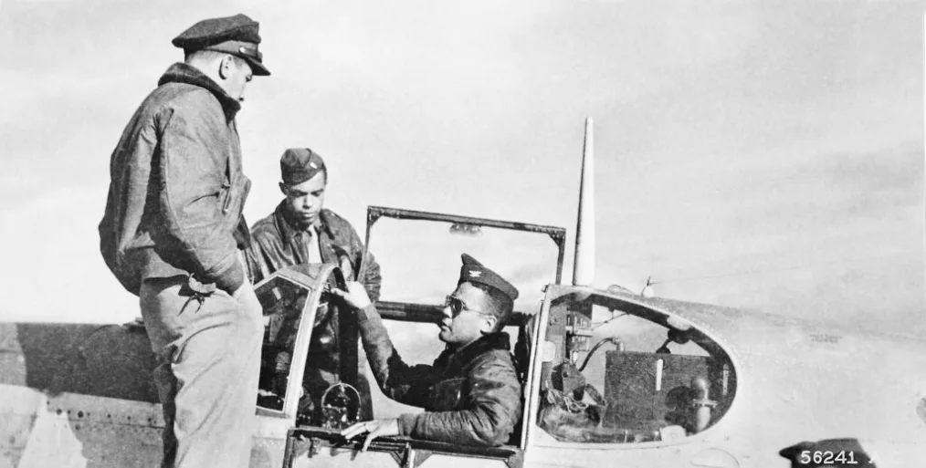 A black and white photo shows three airmen seated in a military truck in conversation. 