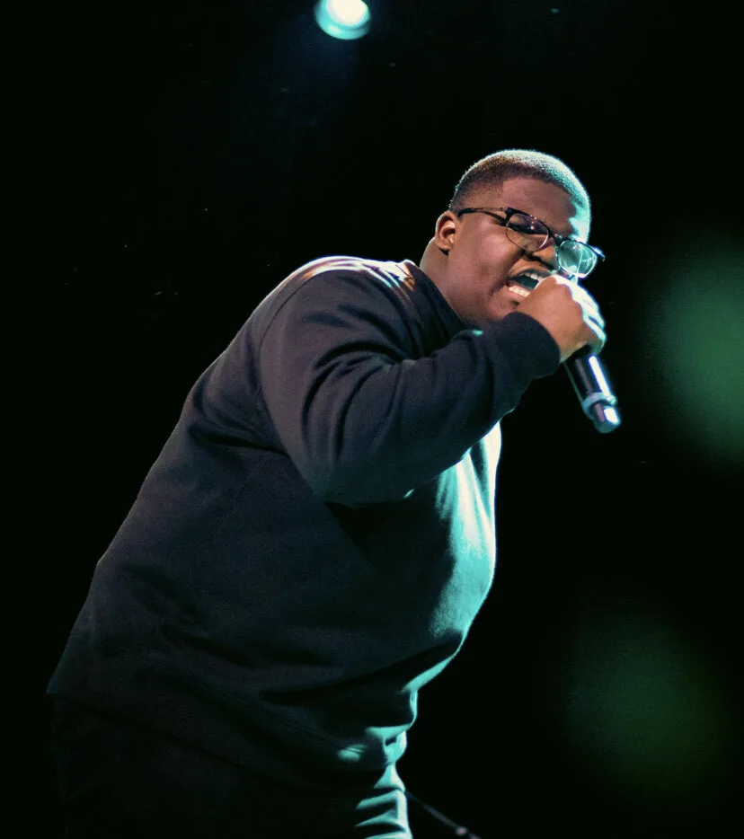 A Black man with short hair and glasses clutches a microphone and appears to be singing or speaking into it. His eyes are closed. Behind him are green and blue out-of-focus lights.
