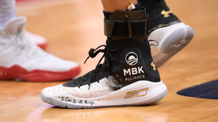 A yellow, white, and black Stephen Curry Signature Under Armour shoe is worn on a basketball court. The person is wearing socks with a white Obama Foundation rising sun logo with the words, "MBK Alliance" on it.