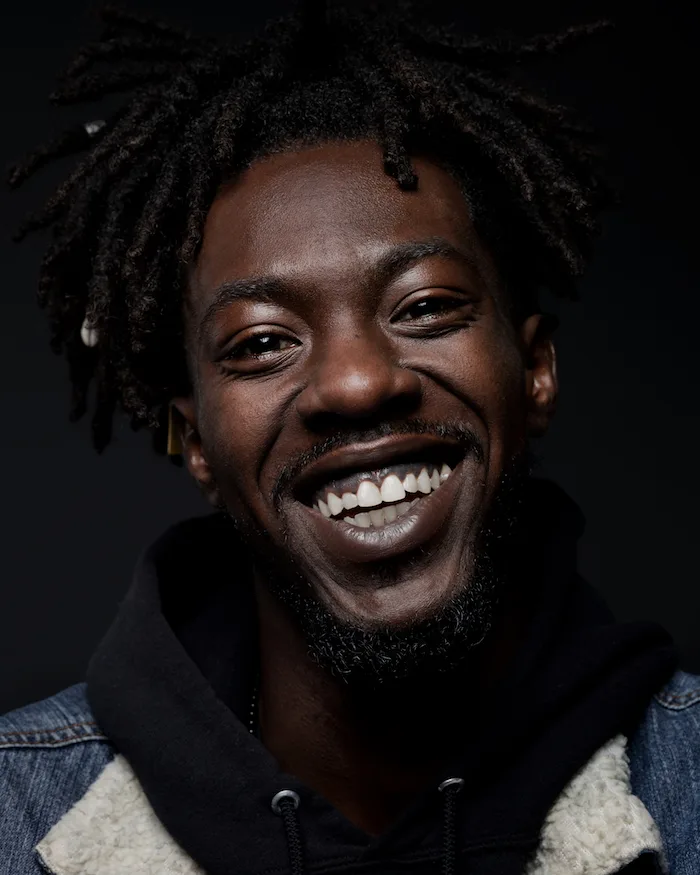 A man with a deep skin tone wearing a black hoodie, denim jacket, and locked hair, smiles in front of a gradient black background. 