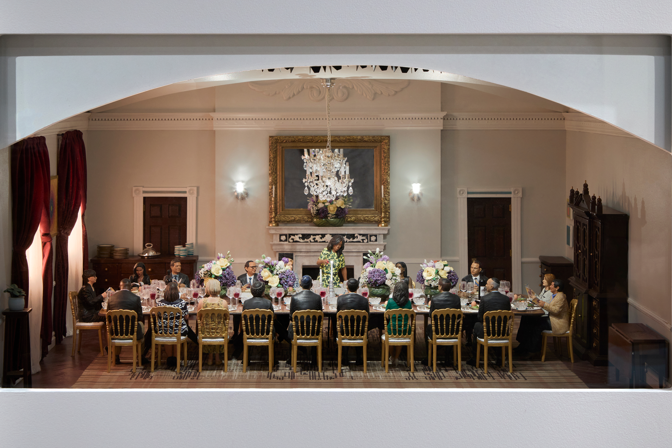 An image of a miniature replica of Mrs. Obama hosting a dinner inside the White House. The miniature features a long and ornate table covered with serviceware and flowers. There are people sitting at the table. Mrs. Obama is standing up at the center of the table. 