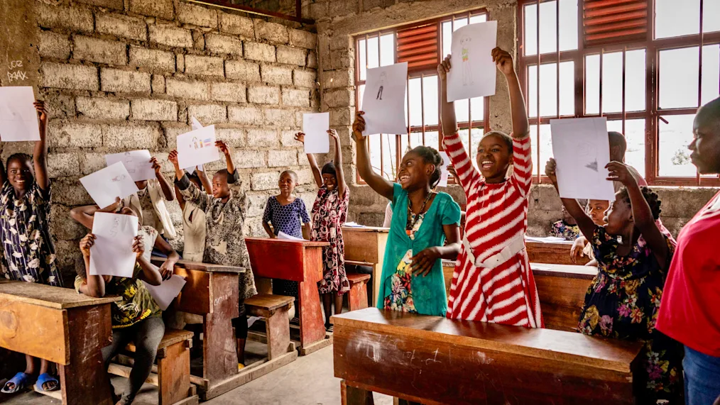 A group of young girls with a range of medium and dark skin tones hold up sheets of paper in a classroom. All girls are standing behind wooden desks. 