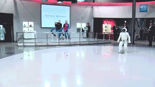 A GIF of President Barack Obama kicking a gray ball back and forth with a Honda Asimo robot that is all white with a black shield over its "face". There are playing on a shiny white floor. There is a crowd watching them.  