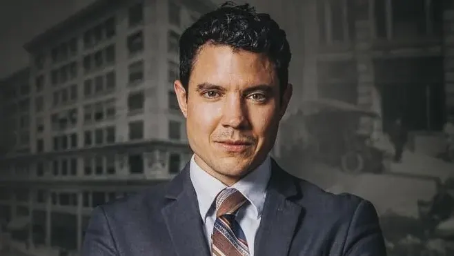 Headshot of Dylan Orr. Dylan has an olive complexion with dark wavy hair. He is wearing a dark gray suit with a striped tie. In the image he is standing with his arms crossed on his chest. He has a wedding ring on his left finger.