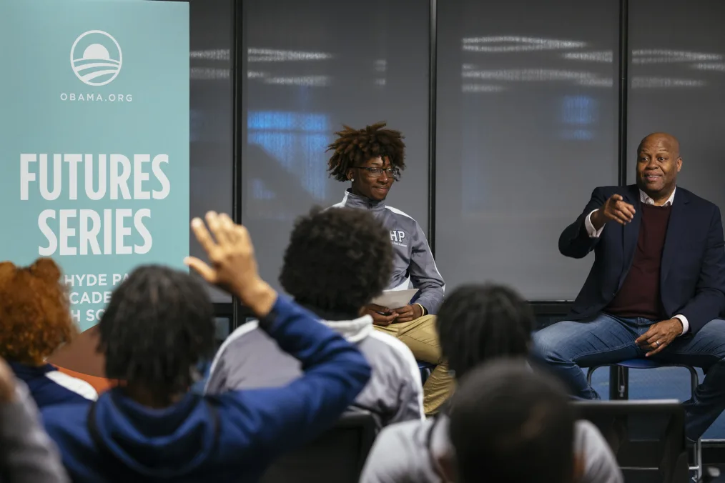 A deep skin toned teenaged boy and a medium-deep skin toned middle-aged man are sitting facing a crowd of people. The boy wears glasses and is dressed in chino pants and a gray zip up jacket. The man is wearing a blazer, blue jeans and a red sweater. Behind them is a light teal, partially obscured sign that reads OBAMA.ORG FUTURES SERIES