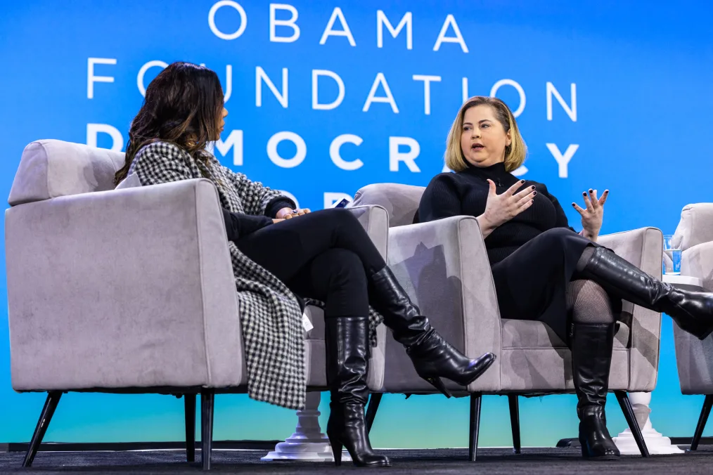 Mandy Teefey, a woman with a light skin tone and short blonde hair speaks on a panel alongside Regina Hall, a Black woman with a medium skin tone. The screen behind them reads, “Obama Foundation Democracy Forum.”