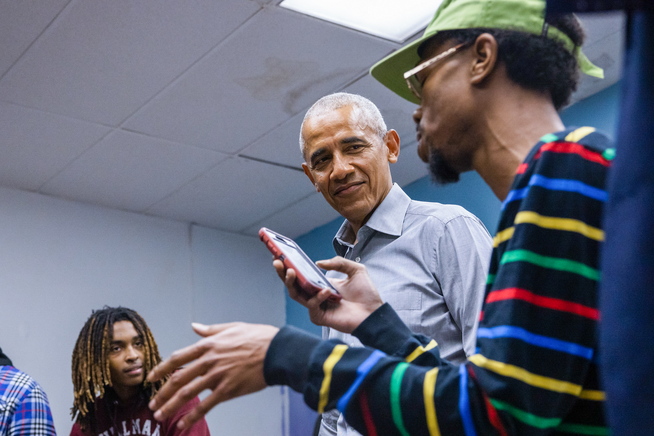 President Obama smiles as he listens to a Black male with a deep skin tone rap from his phone. He is wearing glasses and a neon green cap. Three males an