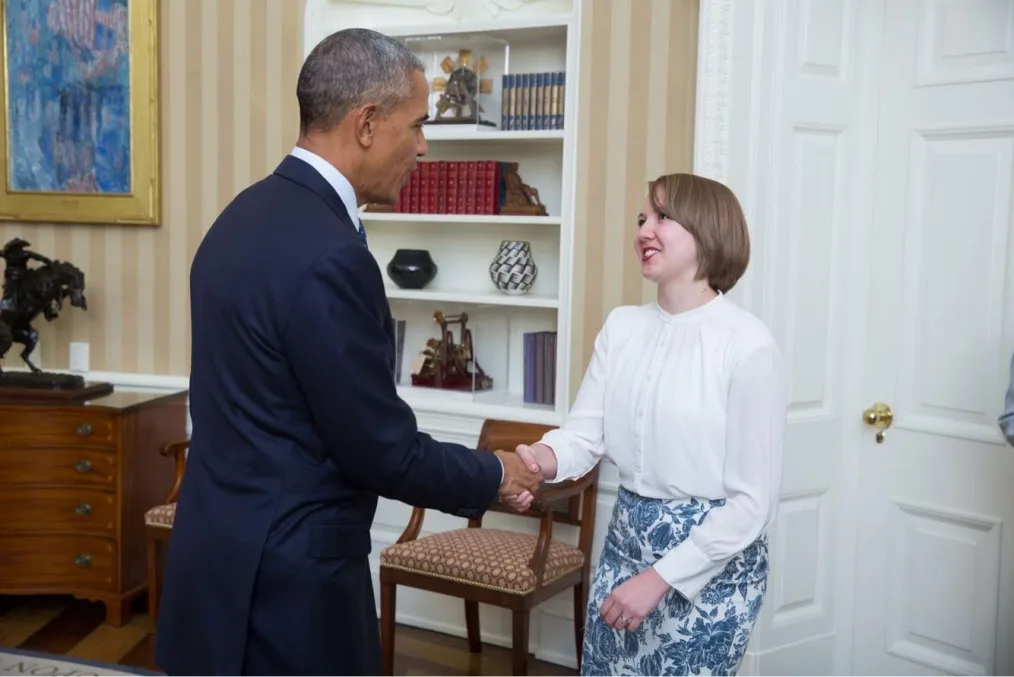 The image is a candid shot of Kolbie Blume shaking hands with President Barack Obama in the Oval Office. The Oval Office has striped tan colored walls and a white bookcase that has a series of red books, blue and gold books, vases, and statues on it. In font of the bookcase is a wooden chair. To the left of the bookcase is a brown wooden desk with a bronze statue sitting on top of it. On the wall is a watercolor painting held in a gold frame. In the photo Kolbie is smiling at President Obama. They have pale skin and light colored hair that falls to their ears. They are wearing a white long sleeved blouse and a blue and white patterned skirt. President Obama has a light medium deep skin complexion and black and grey hair. He is wearing a dark blue suit with a white collared shirt underneath.