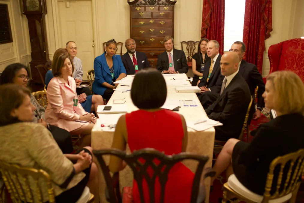 A room of various people mainly light neutral skin tones sitting at a table looking at a woman with a medium brown skin tone wearing a red dress and a short brown bob 