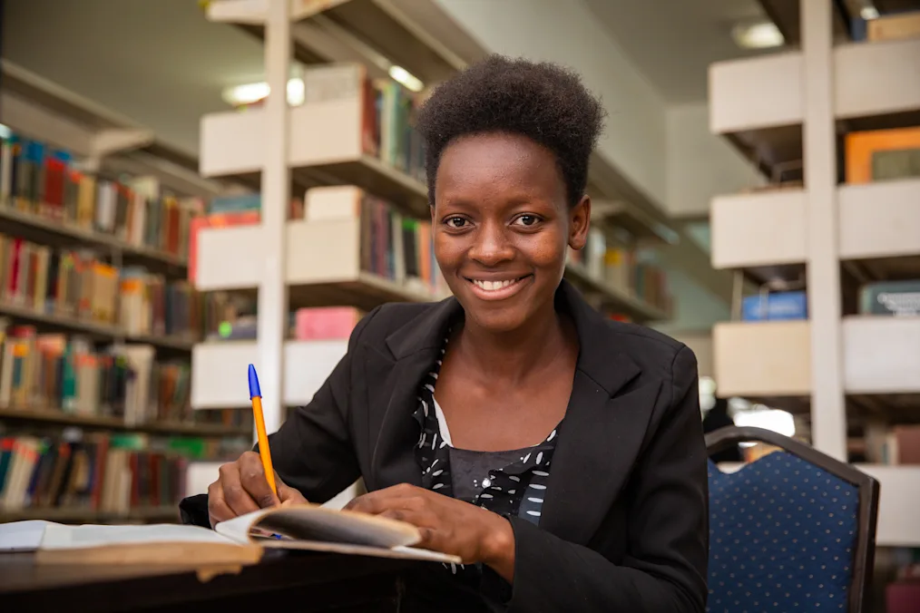 Nimon, a young Kenyan woman, sits at a table with a notebook and pen in hand. She is wearing a black blazer and blouse, with shelves of books pictured behind her. 