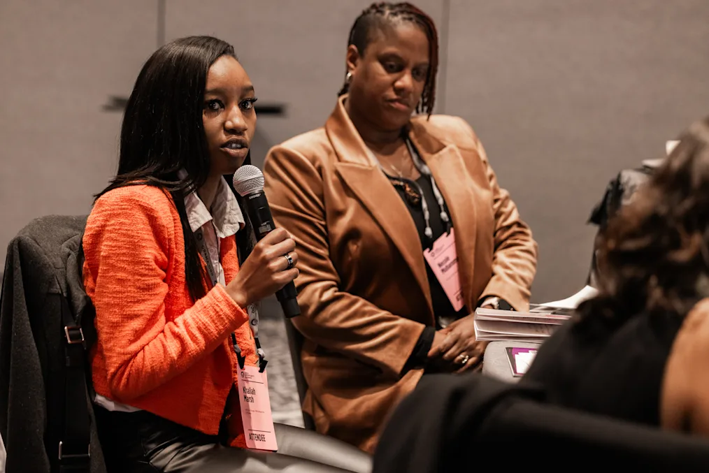 Two women with dark skin tones stand in a room. One woman holds a microphone. Both are dressed professionally. 