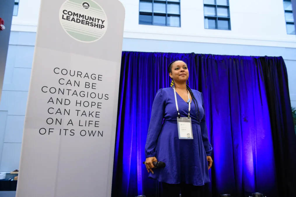 A woman with a medium skin tone and black braids stands smiling in front of a sign that reads:  “ Community Leadership: Courage Can Be Courageous And Hope Can Take Own A Life Of Its Own”