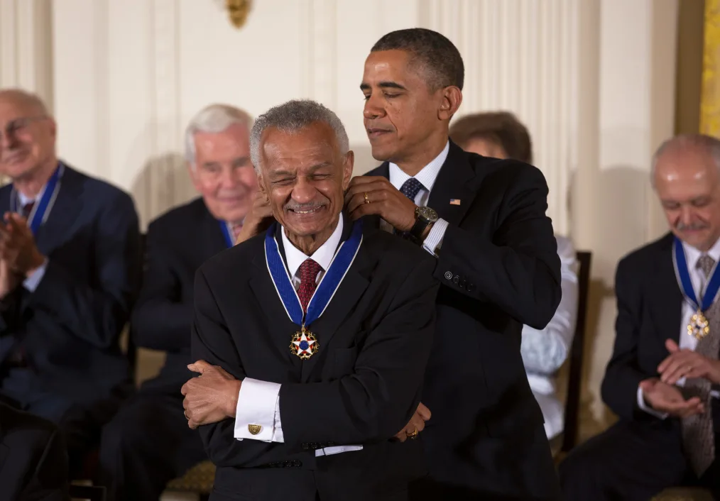 Barack Obama a black man wearing a black suit putting a medal around another man with warm golden brown skin tone smiling and wearing a suit, Three men and one woman with light neutral skin tones sit behind them clapping 