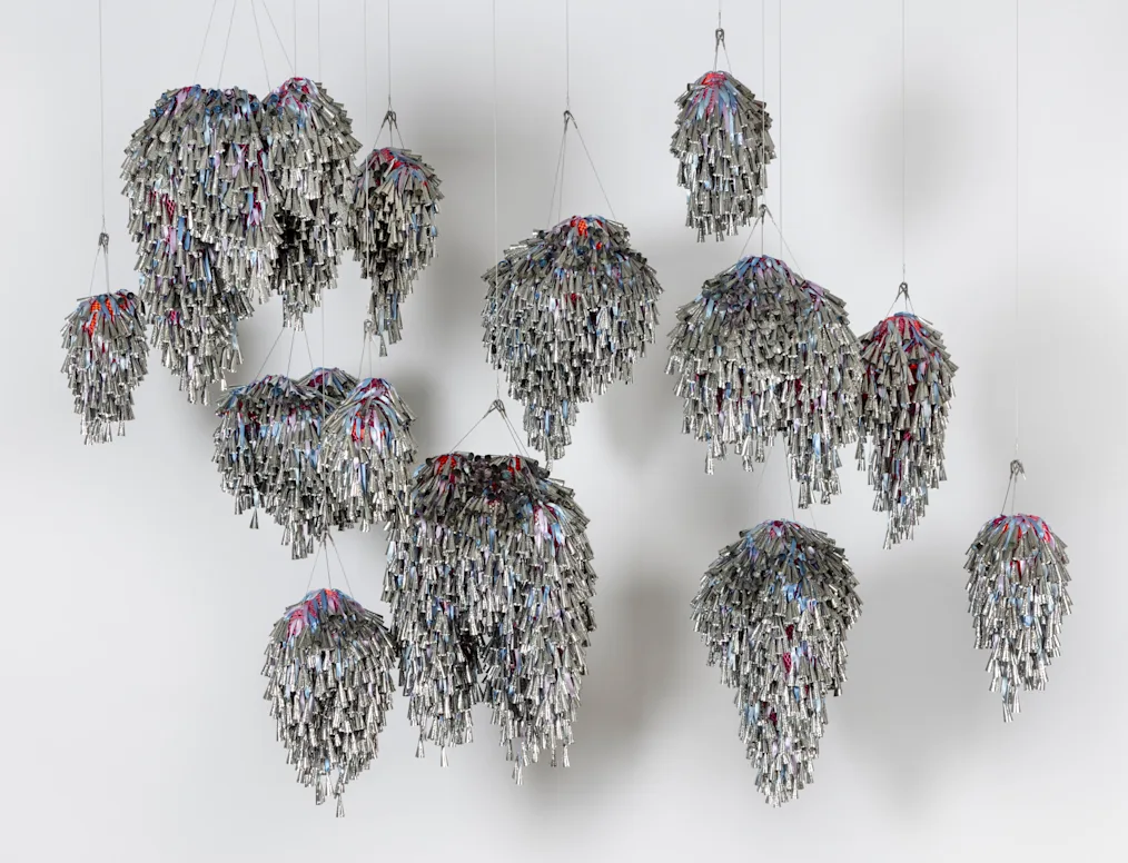 An image of Marie Watt's installation, "Thirteen Moons." The image features an installation that is made up of tin jingles hanging from the ceiling. The jingles are silver cones. There are thousands of them. In between the jingles are straps of pink and blue fabric.