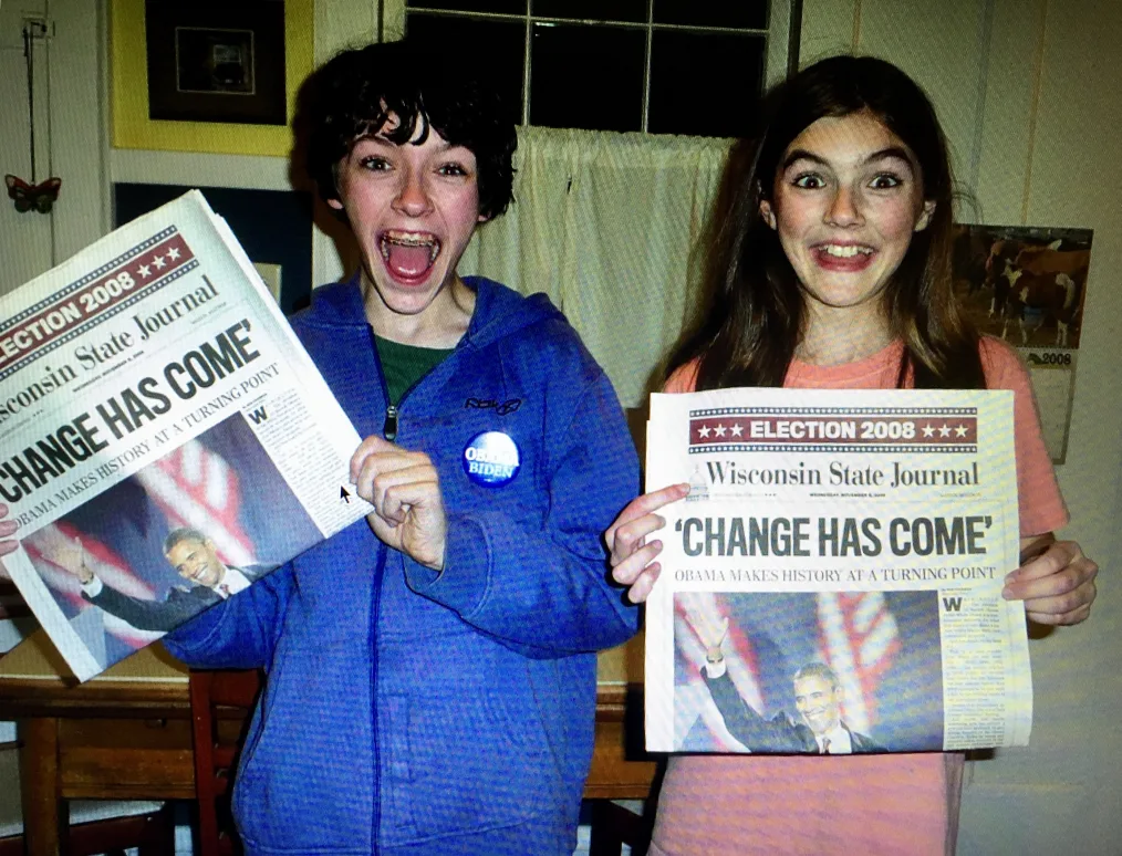 Two excited-looking teenagers hold up newspapers that read "Election 2008, Wisconsin State Journal, "Change Has Come." One wears braces and a jacket with a button reading "Obama Biden."