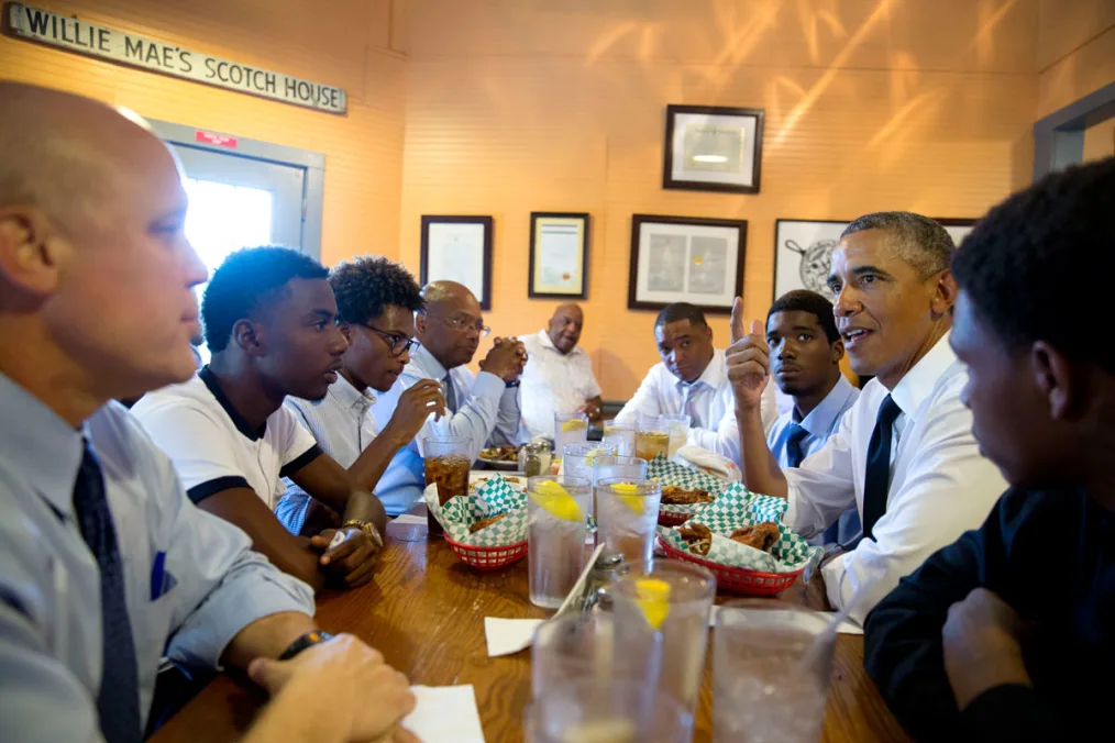 Barack Obama sits at a table speaking with Black men, who look in his direction. On the table in front of them appears fried food in red lattice baskets and water cups with ice and lemon slices. Behind them is a sign that reads "Willie Mae's Scotch House."
