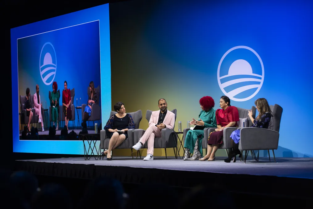 A group of four people sitting down on gray chairs with small microphones  
connected to their ears, appear to be engaging in a meeting together.
The Obama Foundation logo appears on two screens on a blue background
 in white text.