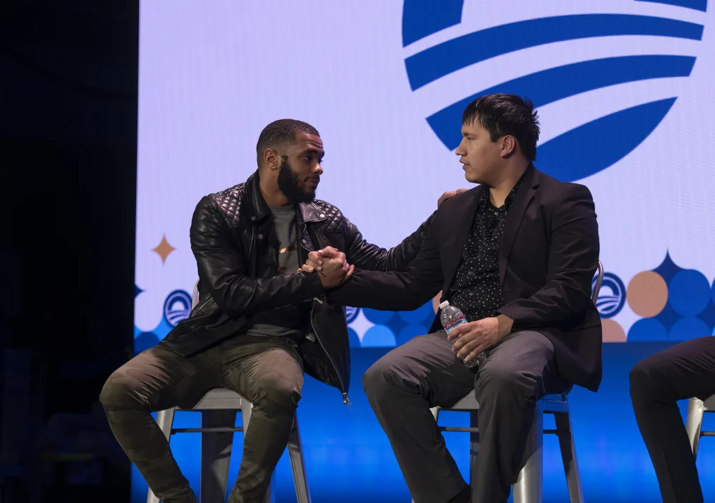 A man with a medium-deep skin complexion is leaning over and holding the hand of another man, who has a medium skin complexion, while touching his shoulder with the other hand. Both individuals are sitting on stools in front of a screening of the Obama Foundation logo. 