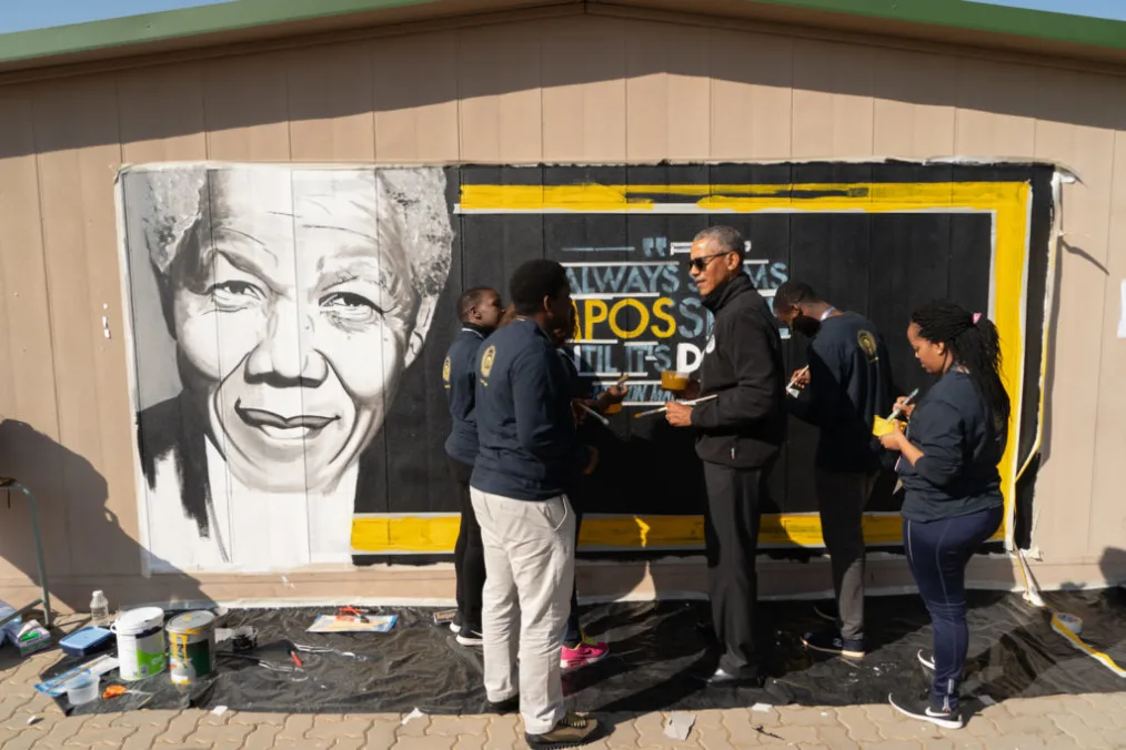 President Obama stands outdoors with sunglasses with a group of men and women ranging from medium to deep skin tones as they paint a mural on a wall with yellow, blue, and white paint