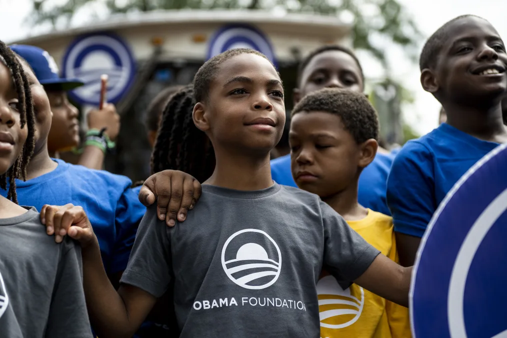 A young boy with a medium-deep skin tone stands proudly in a crowd of other young boys with medium-deep and deep skin tones. He wears a dark gray t-shirt with the Obama rising-sun logo and has his hands on the shoulders of those next to him while another puts their hand on his shoulder.