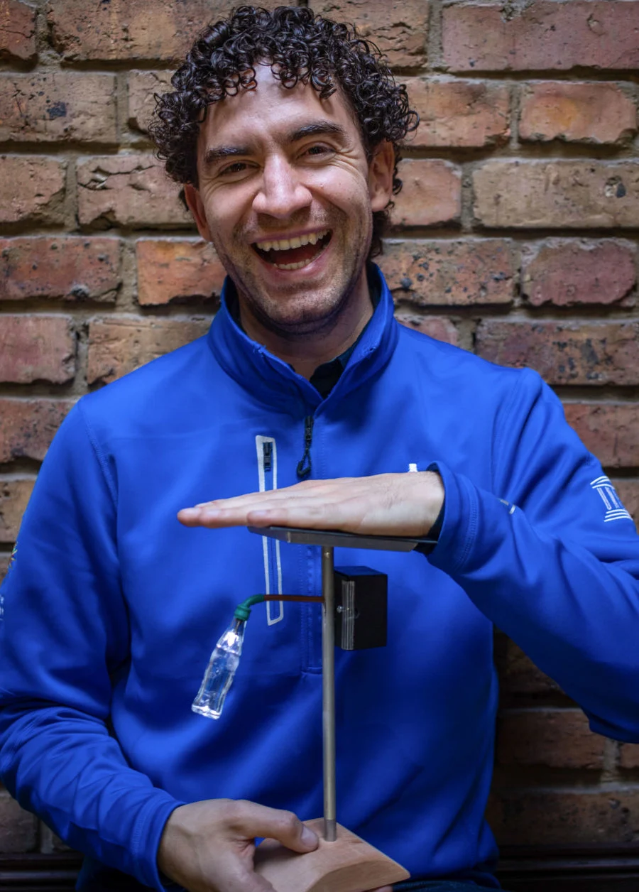 A man with a light skin tone smiles as he holds an electrical creation. He has short black curly hair and is wearing a blue zip-up jacket.