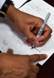 There is a hand with a medium-deep skin tone writing on a sheet of paper using a thin black sharpie marker. The text on the paper that stands out is 