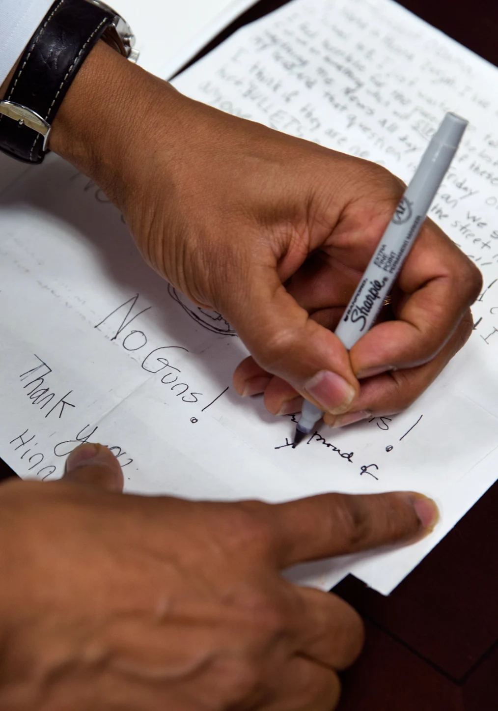 There is a hand with a medium-deep skin tone writing on a sheet of paper using a thin black sharpie marker. The text on the paper that stands out is "No Guns!"
