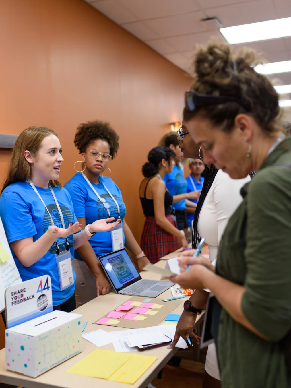 A woman with a light skin tone, a blue shirt with black letters, and an identification tag on a lanyard around her neck stands next to a woman with a medium-deep skin tone, a blue shirt with black letters, and an identification tag on a lanyard around her neck, as they both stand behind a long rectangle table. On the other side of the table, in front of the two women, are two other women. One woman has a medium-deep skin tone with glasses and a white shirt, and the other woman has a light-medium skin tone with a green sweater. There are others blurred out on the ends of both sides of the table. 