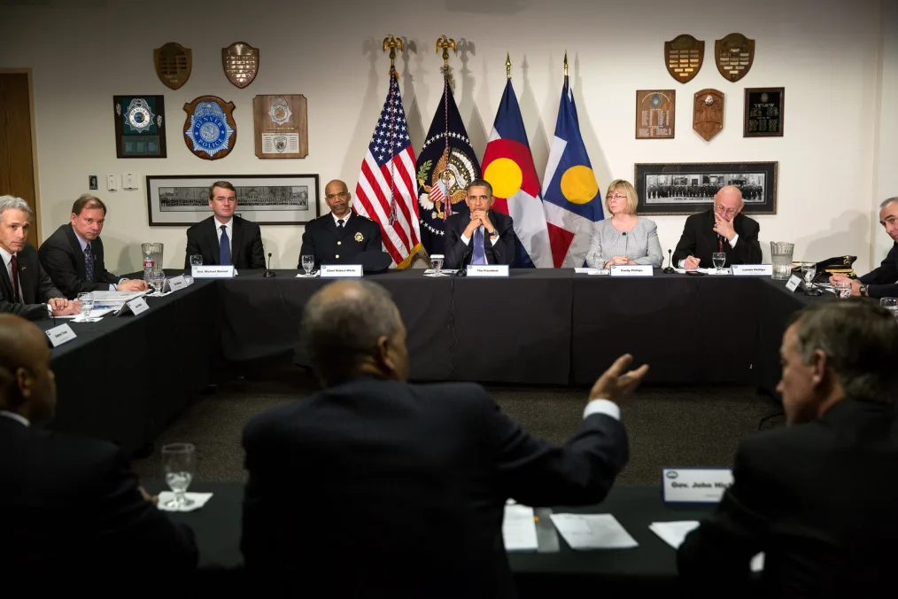 President Obama sits at a square black table with space in the middle. There are a group of individuals around him with different skin tones. There is a white wall behind him with with multiple plaques and photos on it and four flags in front of it. 