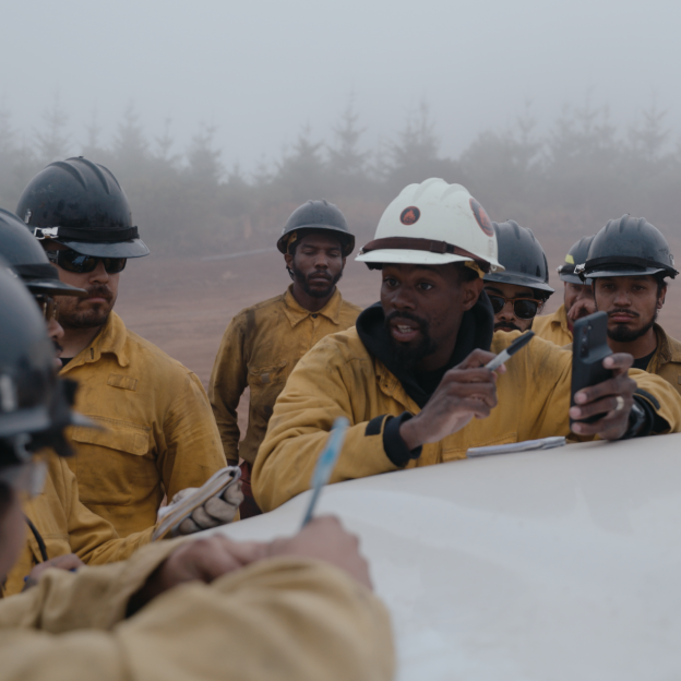 Royal Ramey, a Black man with a dark skin tone, stands between a group of men with a range of light to dark skin tones. They are outdoors and dressed in firefighter protective gear and black hard hats. Ramey is wearing a white hard hat.